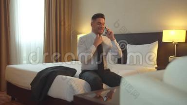 穿着衬衫和领带的人坐在酒店的床上，打电话，脱鞋